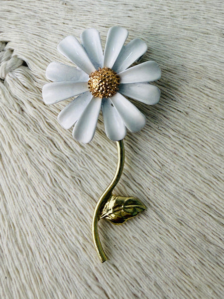 A silver brooch designed as a daisy with a white flower head, a gold-stated center, and a gold stem with a leaf on it, broached onto a textured white surface.
