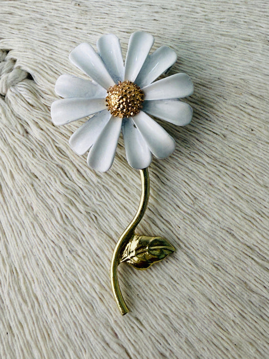 A silver brooch designed as a daisy with a white flower head, a gold-stated center, and a gold stem with a leaf on it, broached onto a textured white surface.