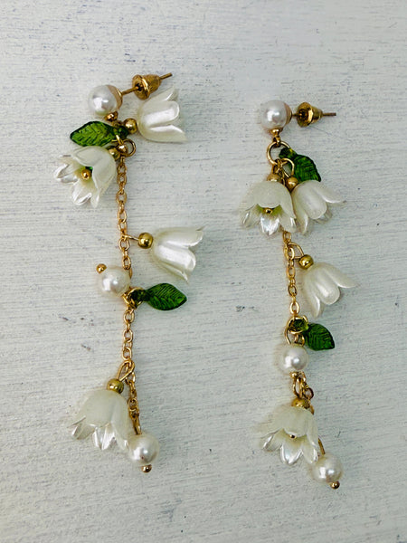 A pair of earrings featuring white lily flowers, green leaves, and gold-colored accents.
