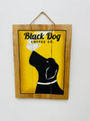Hand-painted Black Dog wooden sign with yellow background, dog and coffee cup illustration, and hanging rope on white background.