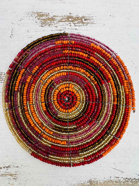A round beaded coaster with orange, red and gold colors, displayed on a white surface.