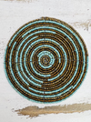 A circular beaded coaster with a turquoise and gold color pattern, placed on a wooden surface.