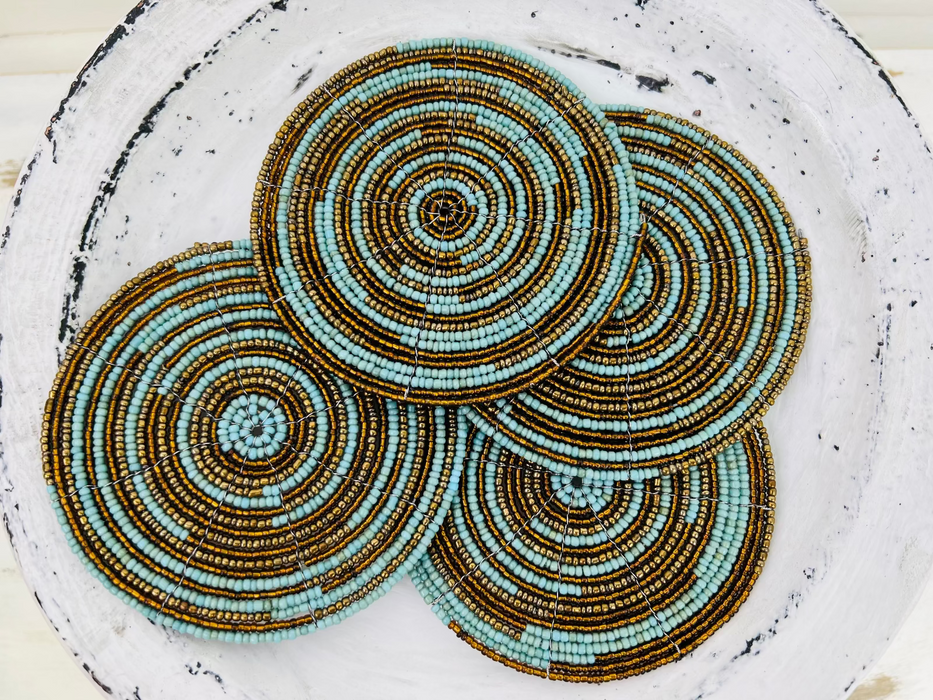 Set of turquoise, gold and olive beaded coasters arranged on a light wooden surface.