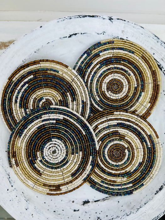 Set of handmade beaded coasters in natural white, gold and paua-blue colours, arranged on a light wooden background.