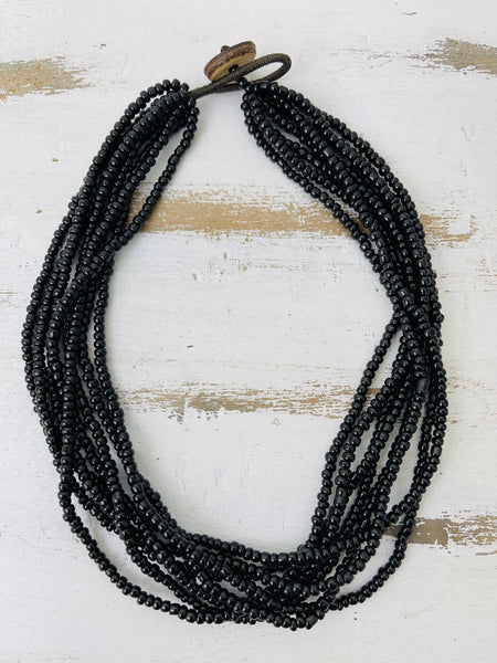 A multi-strand black beaded necklace with a dark metal clasp, displayed on a distressed white wooden surface.