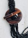 A bohemian-style multi-strand necklace featuring black seed beads, a large circular wooden medallion, and a wooden toggle clasp, shown against a textured off-white background.