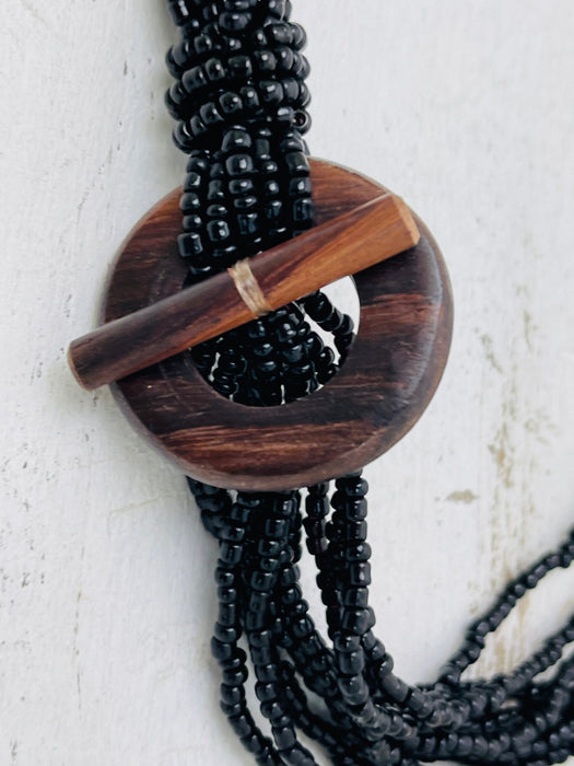 A bohemian-style multi-strand necklace featuring black seed beads, a large circular wooden medallion, and a wooden toggle clasp, shown against a textured off-white background.
