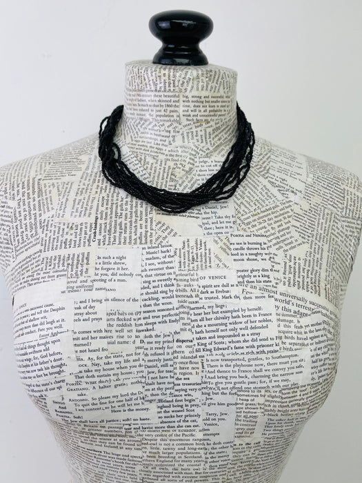 A black multi-strand beaded necklace displayed on a book-print mannequin torso against a plain white background.