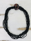 A black beaded necklace with a hand-carved wood circle and T bar closure, displayed on a white wooden background.