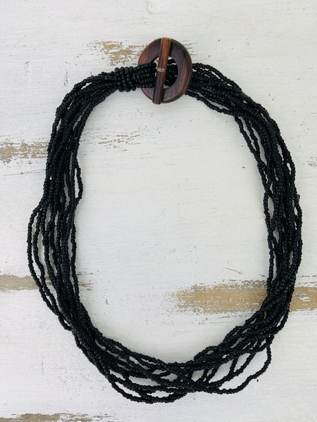 A black beaded necklace with a hand-carved wood circle and T bar closure, displayed on a white wooden background.