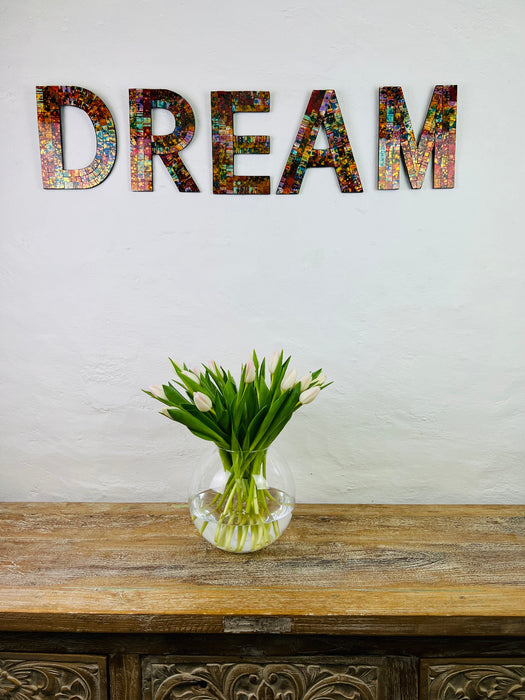 Hand painted glass letters spelling out 'DREAM' mounted on a wall above a wooden table with a vase of flowers on it.