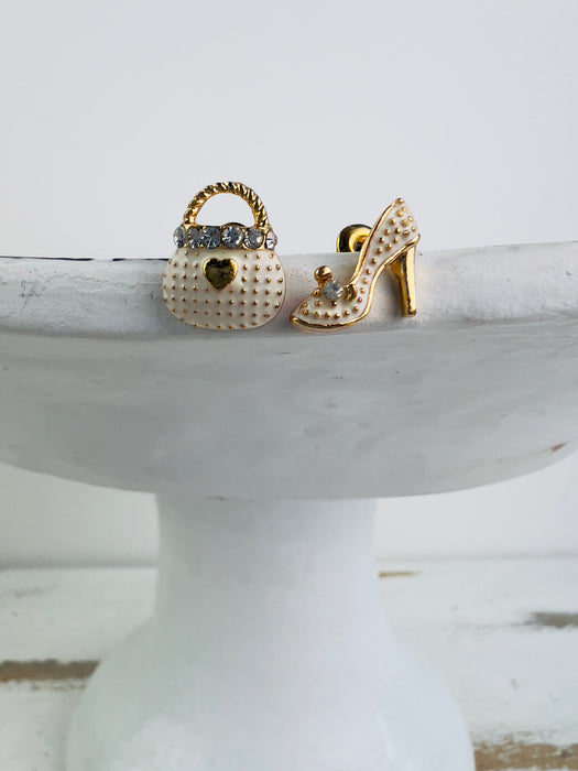 A pair of stud earrings featuring one in the shape of a high heel shoe and the other in the shape of a handbag, both detailed with crystals.