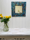 display front view of mosaic square mirror hung on the wall with vase of roses below on table