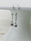 Silver earrings with star-shaped charms hanging on a white hook against a white background.