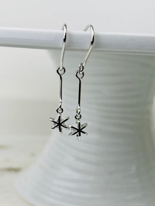 Silver earrings with star-shaped charms hanging on a white hook against a white background.