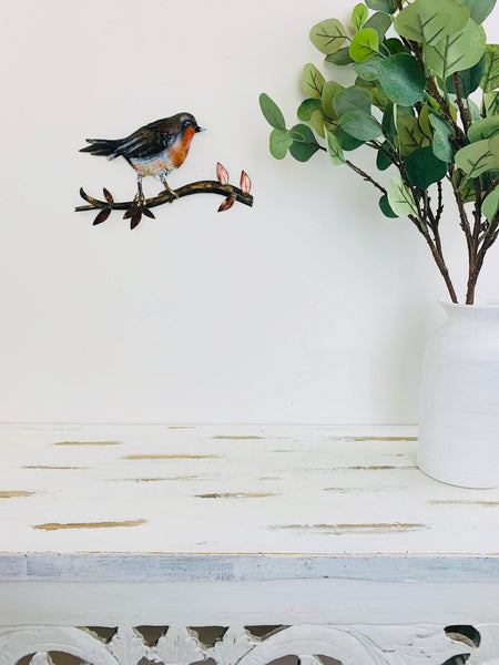 Hand-painted metal robin on a branch mounted on a wall beside a vase with green leaves
