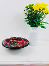 Handmade mosaic glass bowl in Broadway tones on white table beside vase with yellow flowers, showing bright red, blue and yellow mosaic pattern.