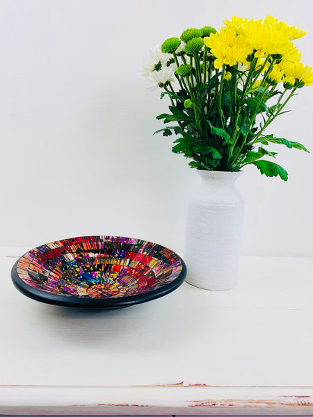 Handmade mosaic glass bowl in Broadway tones on white table beside vase with yellow flowers, showing bright red, blue and yellow mosaic pattern.