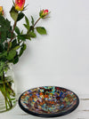 Handmade mosaic glass bowl in Calypso tones on white table beside pink and orange flowers, showing reflective red and gold glass mosaic surface.
