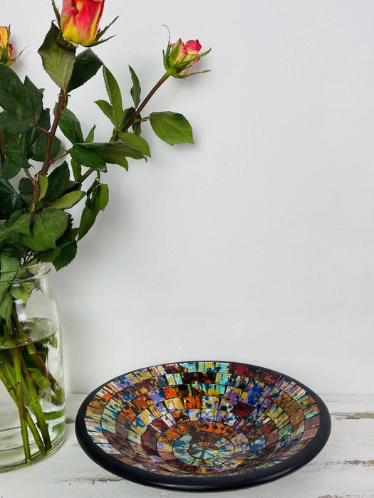 Handmade mosaic glass bowl in Calypso tones on white table beside pink and orange flowers, showing reflective red and gold glass mosaic surface.