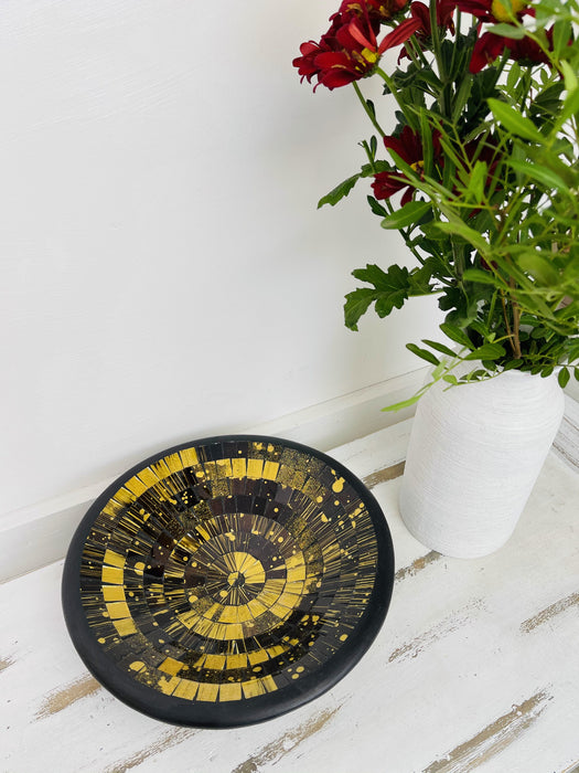 A handmade and hand painted mosaic glass bowl with a terracotta base, featuring a black and gold color pattern, displayed on a white wooden surface with a vase of flowers in the background.