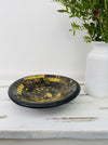 Angled view of handmade mosaic glass bowl in Firecracker gold and black tones on white background beside vase with green leaves.
