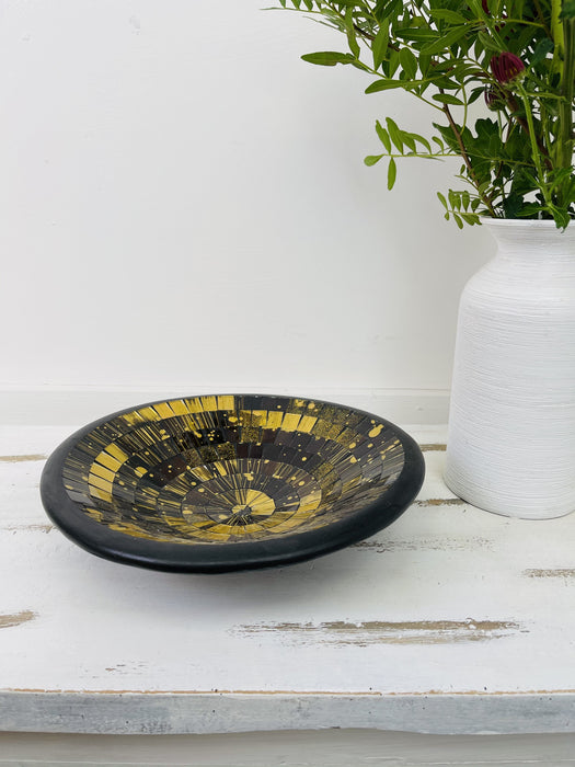 Angled view of handmade mosaic glass bowl in Firecracker gold and black tones on white background beside vase with green leaves.