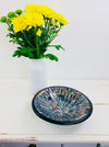 Handmade mosaic glass bowl in Miami tones on white table beside yellow flowers in vase, showing reflective blue, gold and red mosaic.
