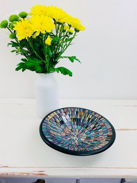 Handmade mosaic glass bowl in Miami tones on white table beside yellow flowers in vase, showing reflective blue, gold and red mosaic.