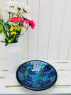 Handmade mosaic glass bowl in Reef tones on white table beside vase with white and pink flowers, showing shimmering deep blue glass mosaic.
