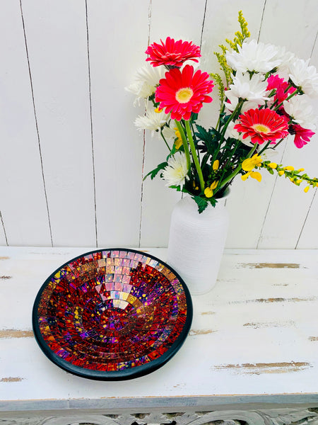 Handmade mosaic glass bowl in Violet Burst tones on white table beside red and white flowers, showing deep purple and red glass mosaic.