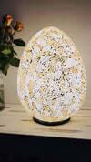 Mosaic egg lamp fully illuminated beside green leaves and orange flowers, giving soft warm glow through gold and silver mosaic glass.