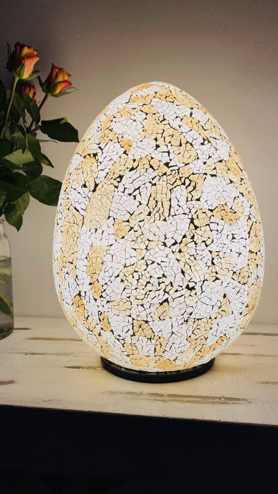 Mosaic egg lamp fully illuminated beside green leaves and orange flowers, giving soft warm glow through gold and silver mosaic glass.
