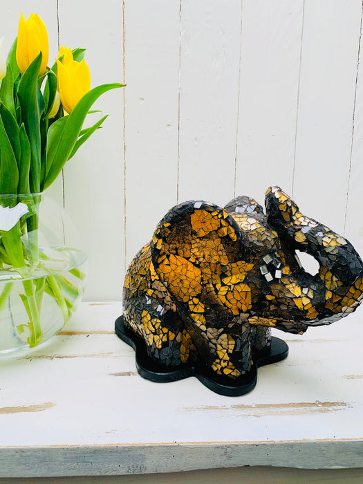 Handmade mosaic elephant lamp illuminated in black and gold tones beside yellow tulips on white surface, showing reflective glass pattern.