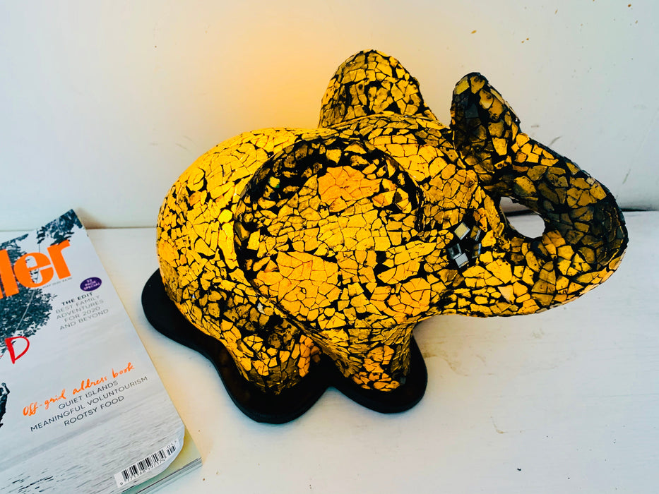 Handmade mosaic elephant lamp illuminated on white wooden surface, glowing softly through gold and black glass mosaic.