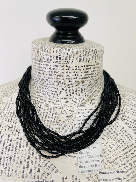 A black beaded necklace draped over a book-page-covered mannequin neck with a black spherical top, photographed against a plain light background.