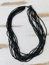 A multi-strand black beaded necklace with a black wood button clasp, displayed on a distressed white and light brown wooden surface.