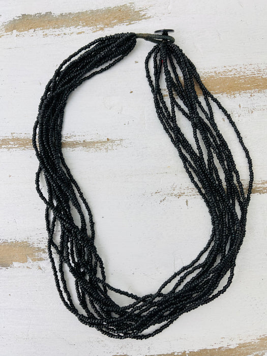 A multi-strand black beaded necklace with a black wood button clasp, displayed on a distressed white and light brown wooden surface.