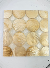 Handmade shell coaster with cream and gold tones, consisting of various circular shells arranged in a pattern on a flat surface.