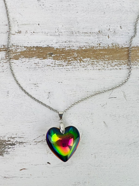 A silver chain necklace with a multicolored glass heart pendant, displayed against a white wooden background.